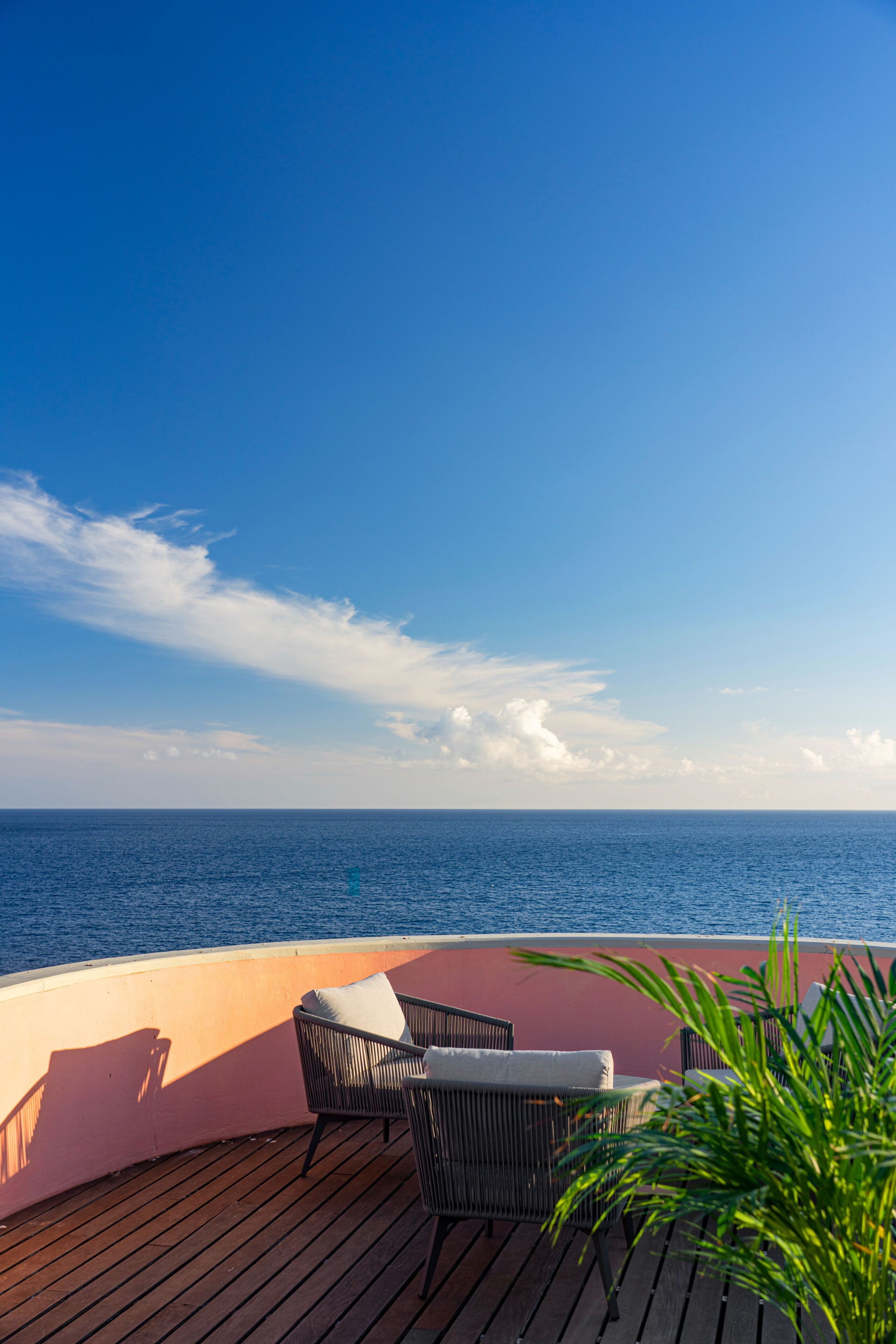 A deck with two wicker chairs and a potted plant, overlooking a calm sea under a clear sky.