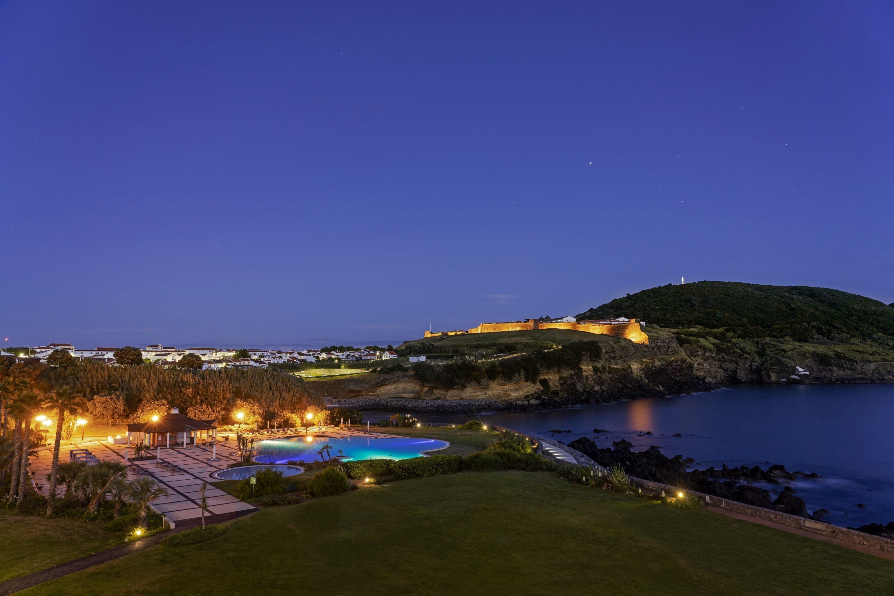 A coastal resort with a swimming pool, a gazebo, and a hilltop fortification.