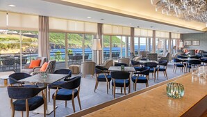 A spacious outdoor dining area with multiple tables and chairs, large windows offering a view of the sea and greenery, and a ceiling with hanging glassware.