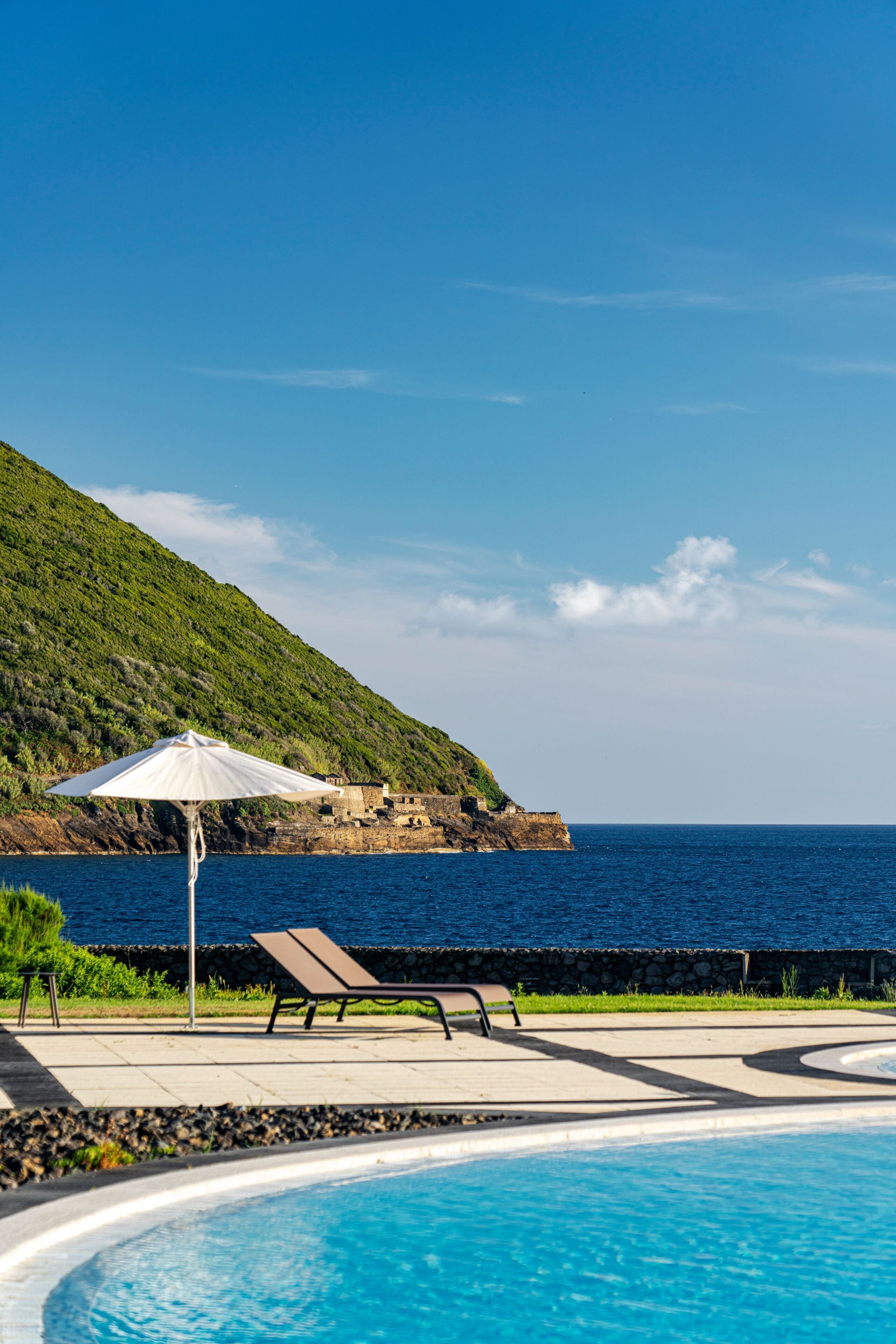 Área de piscina com espreguiçadeiras e um guarda-sol branco, com vista para um corpo de água e uma encosta.