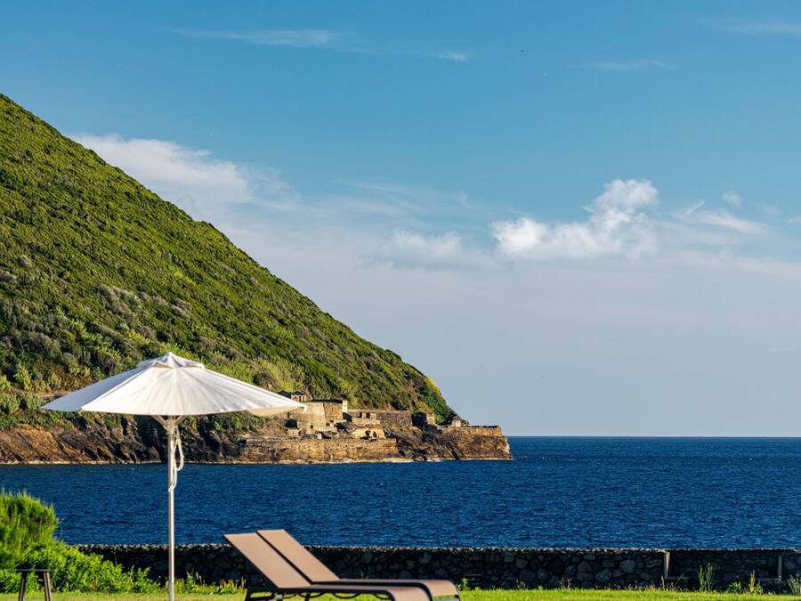 Área de piscina com espreguiçadeiras e um guarda-sol branco, com vista para um corpo de água e uma encosta.