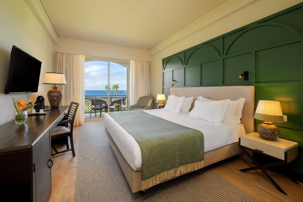 Une chambre d’hôtel avec un grand lit, un bureau, une chaise, une télévision et un balcon avec vue sur l’océan.
