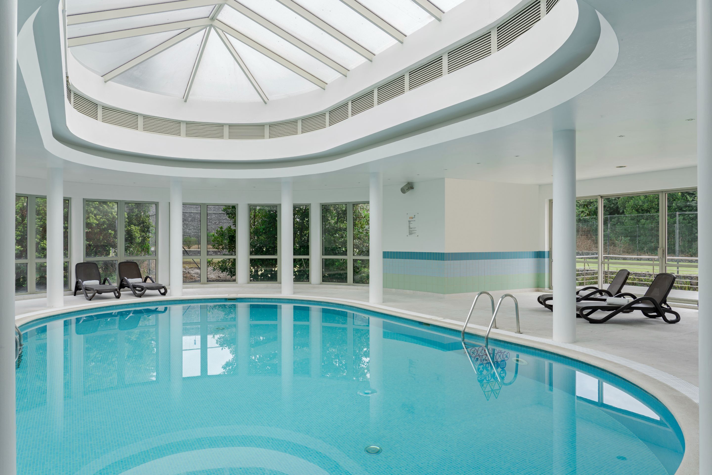 Una piscina interior con techos de vidrio transparente, tumbonas y grandes ventanales.