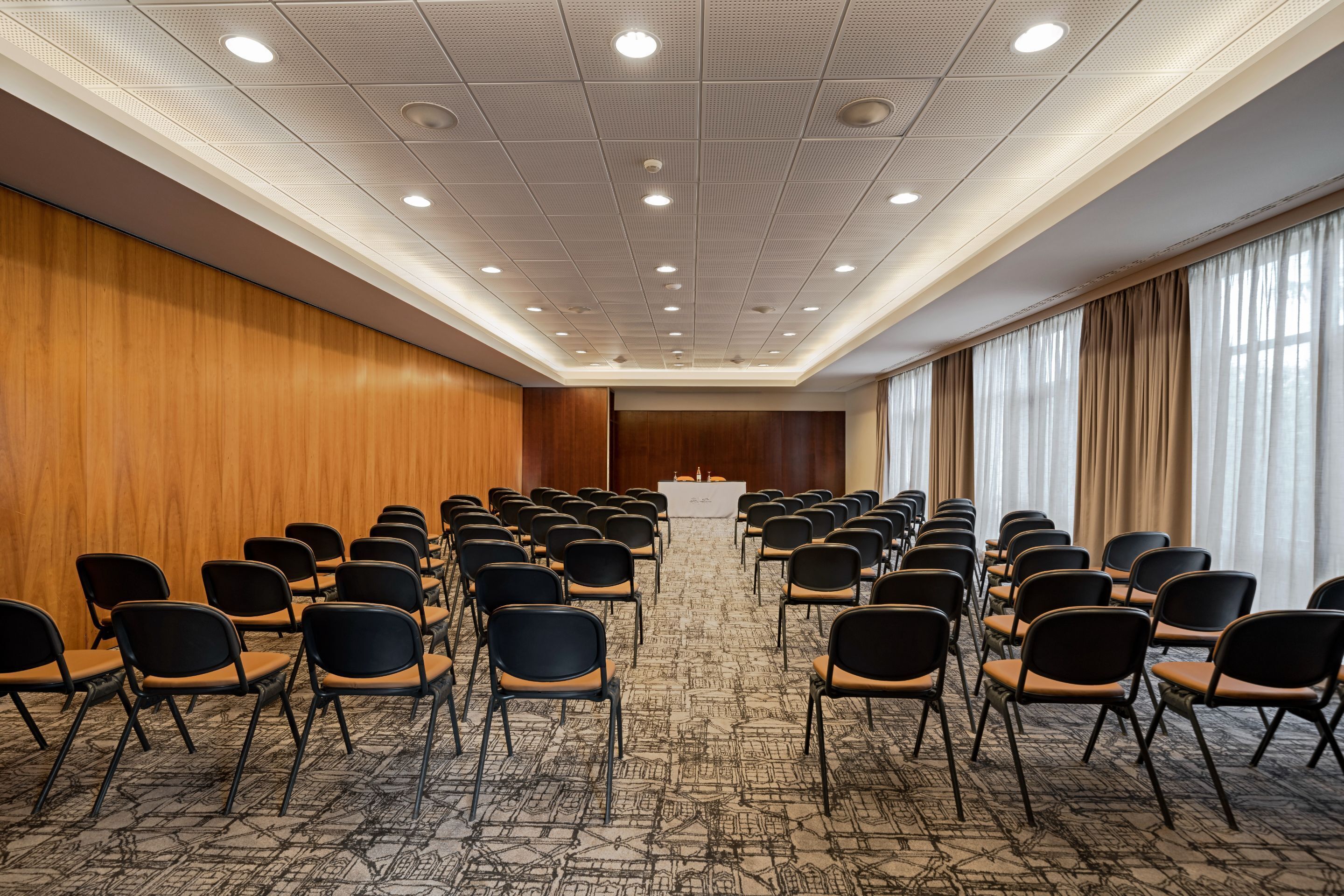 A conference room with rows of chairs facing a podium, wooden panel walls, and large windows with curtains.