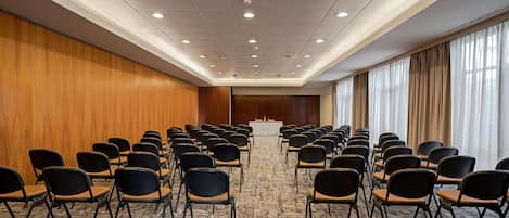A conference room with rows of chairs facing a podium, wooden panel walls, and large windows with curtains.