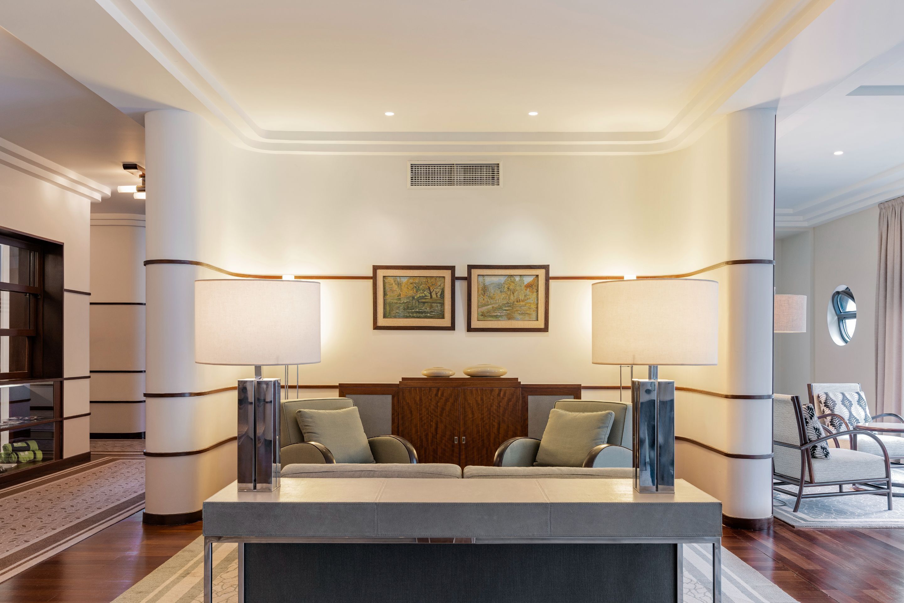 A modern hotel lobby with a sofa set, two framed paintings, a wooden cabinet, and a large mirror.