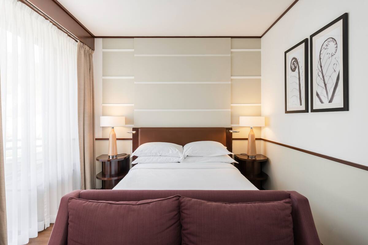 A hotel room with a large bed, two bedside lamps, a headboard with horizontal lines, and two framed pictures on the wall.