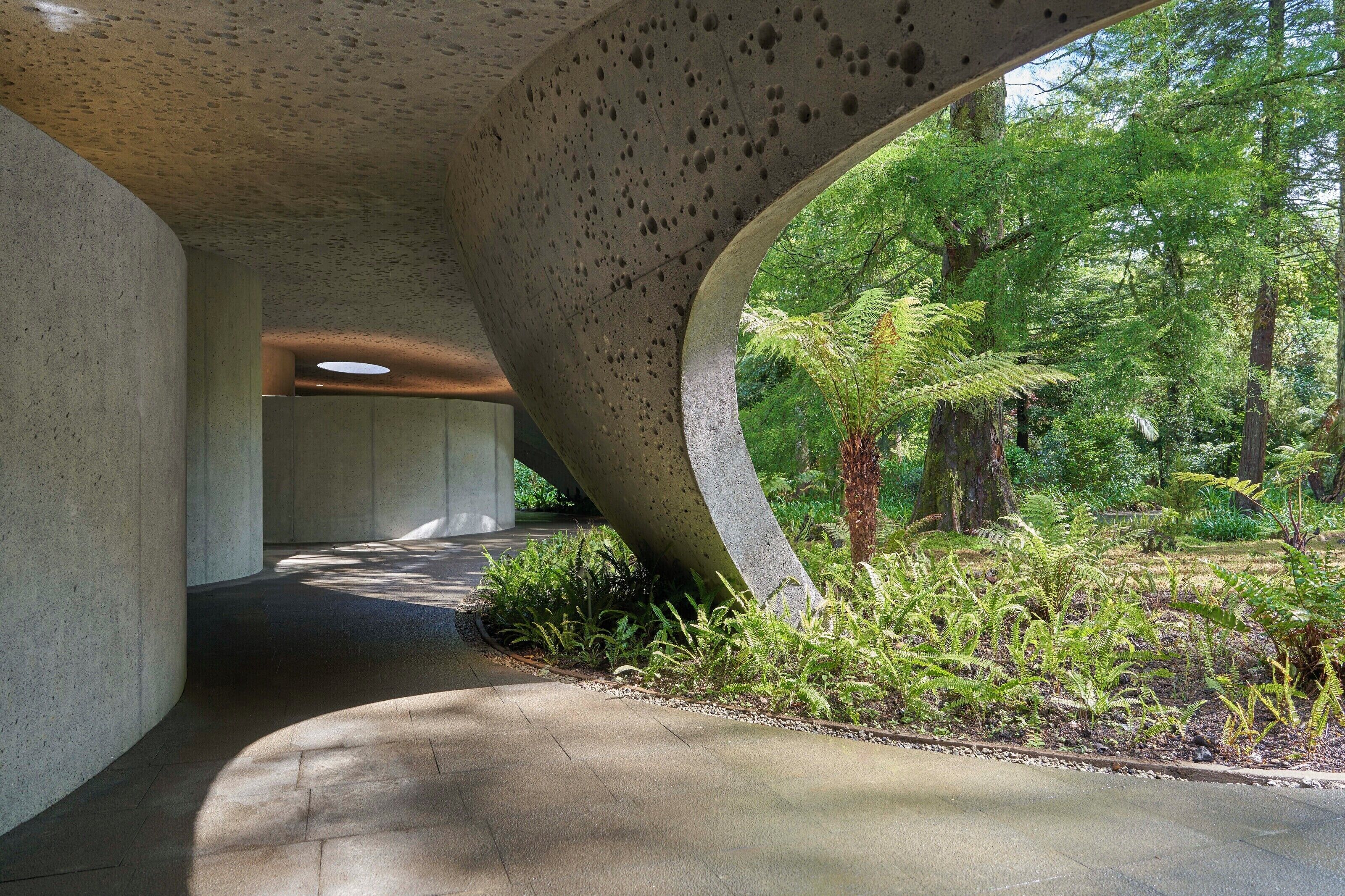 A concrete structure with a curved overhang and a pathway leading through a lush garden.