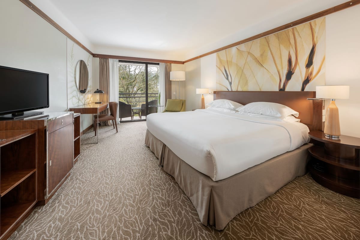 A hotel room with a large bed, a desk with a TV, a chair, and a balcony with a view of trees.
