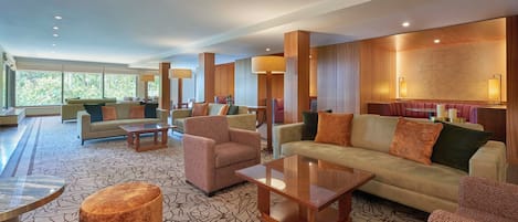 A spacious lobby with multiple seating areas, including sofas and armchairs, arranged around a central coffee table. Large windows provide natural light and a view of greenery outside.