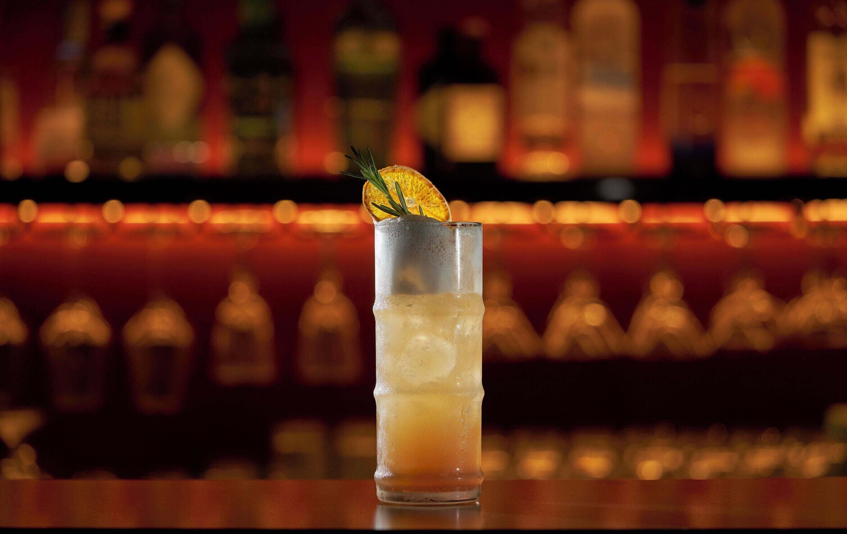 A tall glass with a layered drink, garnished with a slice of orange and a sprig of rosemary, set against a blurred bar background with bottles.