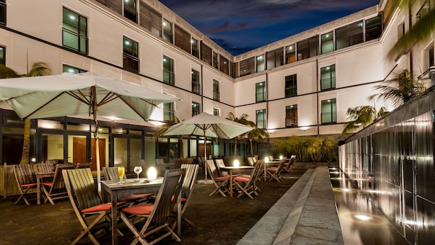 An outdoor dining area with tables and chairs, umbrellas, and a modern building facade at dusk. - Hotel do Canal (Horta)