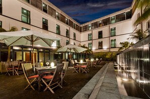 An outdoor dining area with tables and chairs, umbrellas, and a modern building facade at dusk. - Hotel do Canal (Horta)