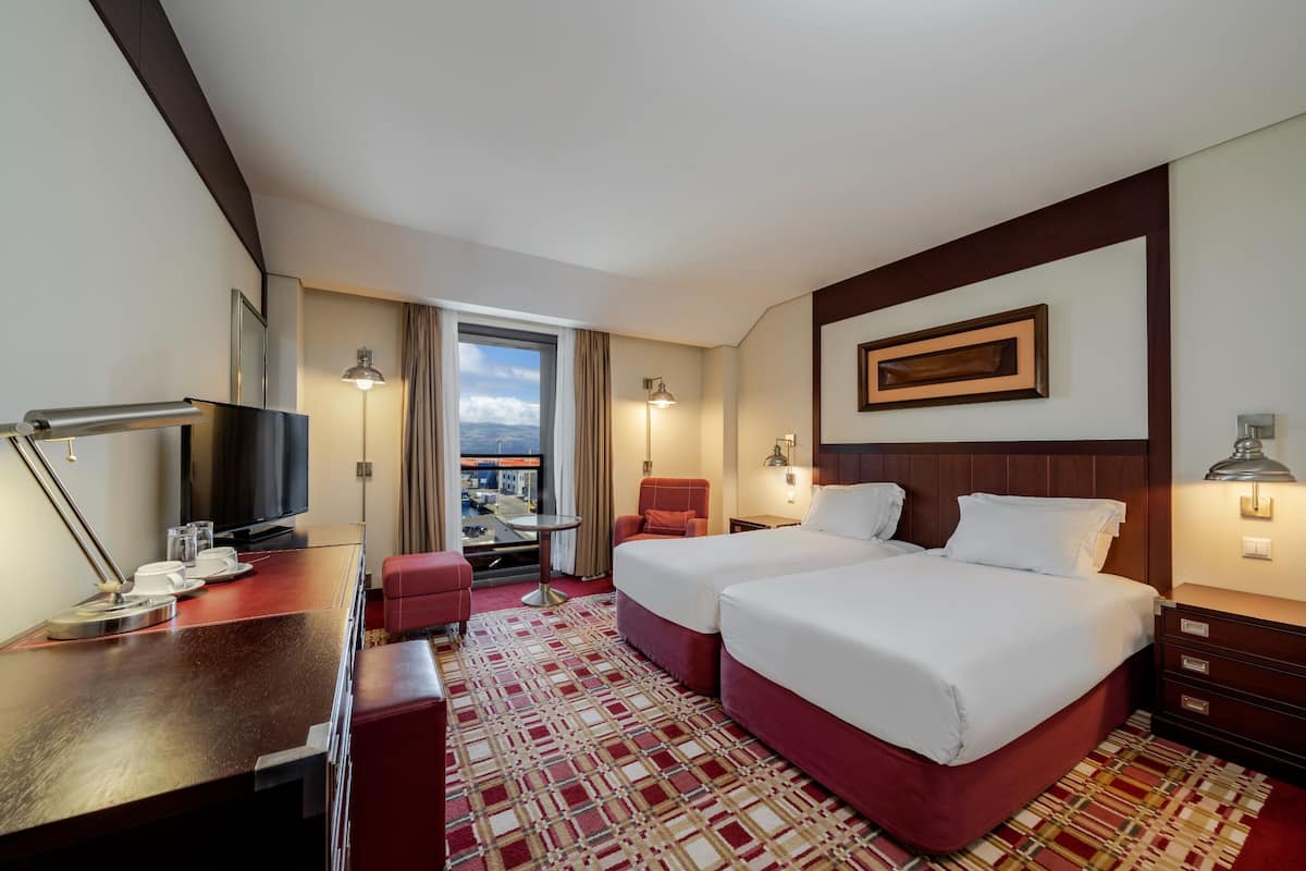 A hotel room with two beds, a desk with a TV, a chair, and a view of the city.