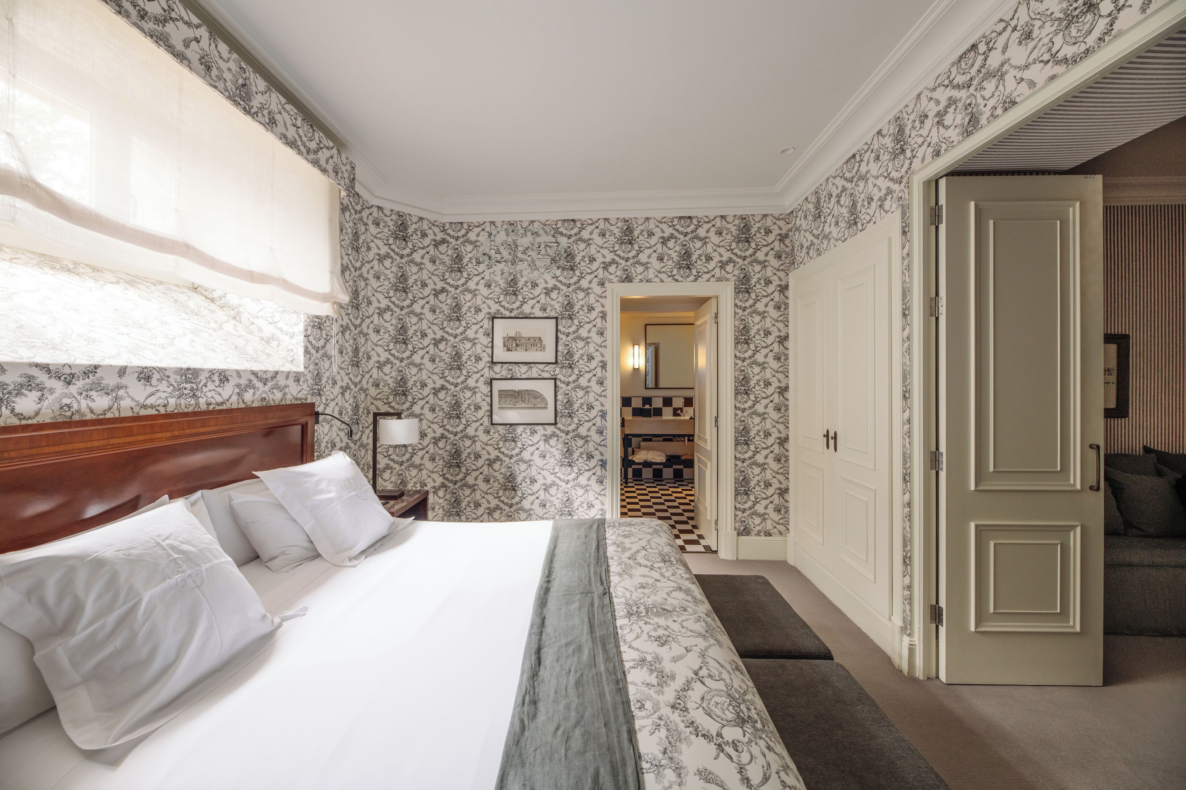 Habitación de hotel con cama, mesita de noche, lámpara, puerta y pasillo que da a un baño.