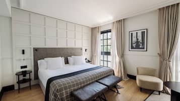Une chambre d’hôtel avec un grand lit, une banquette, une chaise, une petite table et une fenêtre avec des rideaux.
