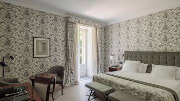 A hotel room with a bed, a desk with a chair, a bench, a framed picture on the wall, and a window with curtains.
