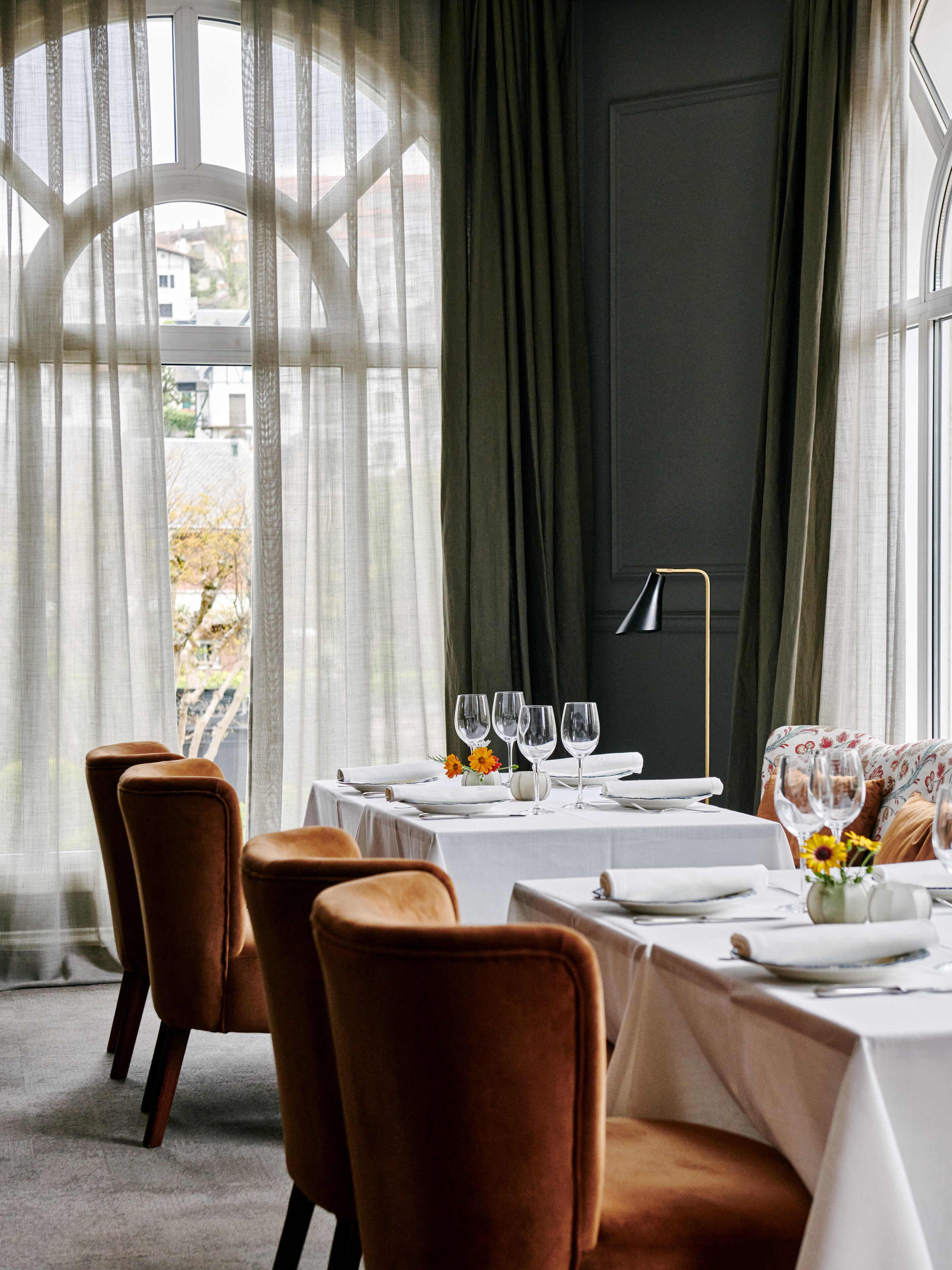 A dining area with a table set for four, chairs, and a view through a window with sheer curtains.