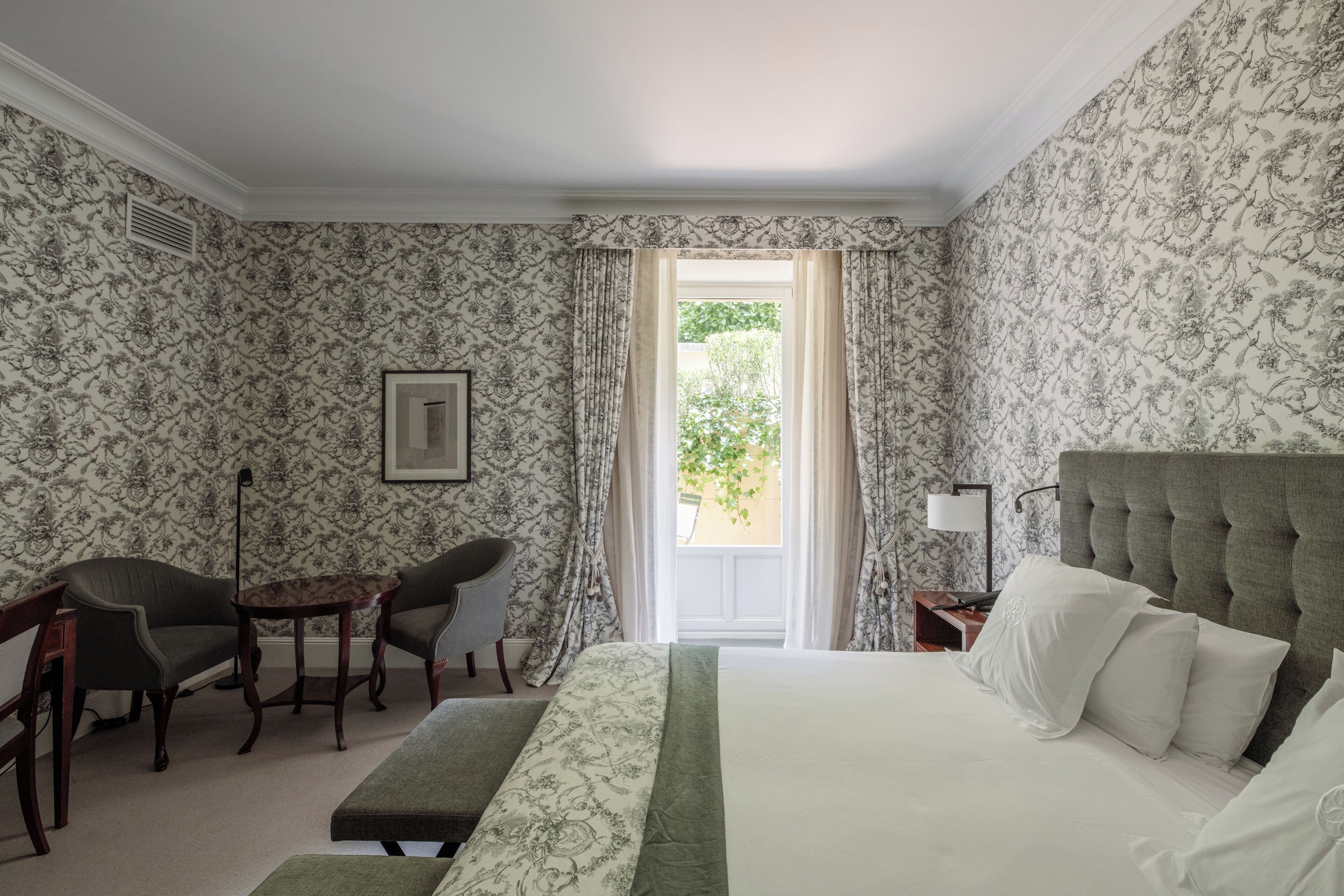 Una habitación de hotel con cama, dos sillas, un escritorio, una mesita de noche y una ventana con cortinas.