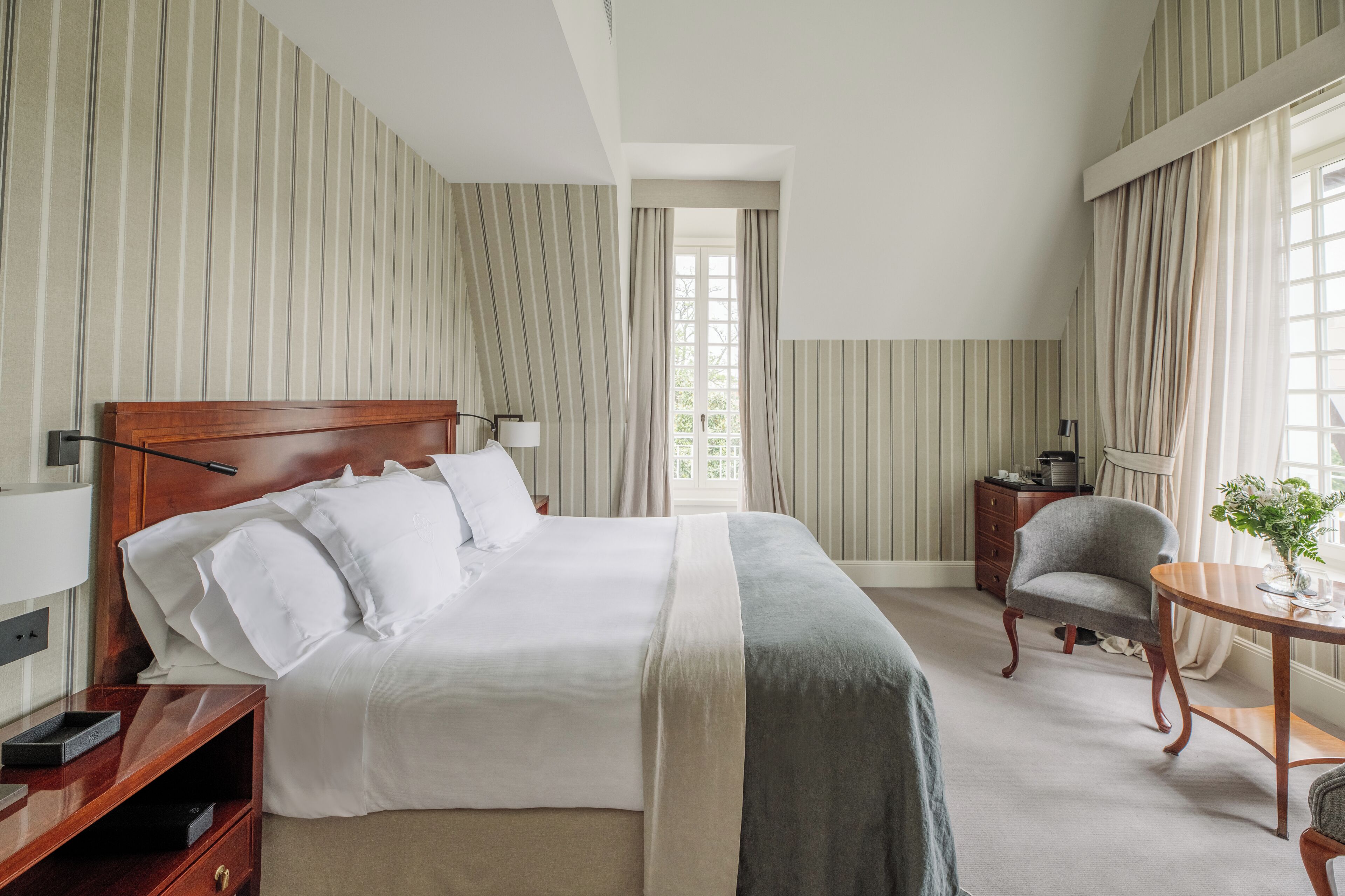 A hotel room with a large bed, a bedside table, a chair, a small table with a vase, and a window with curtains.
