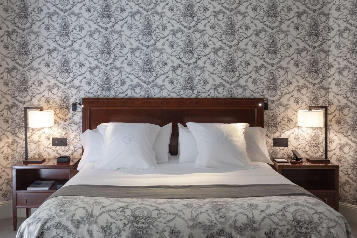 A neatly made bed with white linens and a patterned duvet, flanked by two bedside tables with lamps and a telephone. The room features a wooden headboard and a patterned wallpaper.