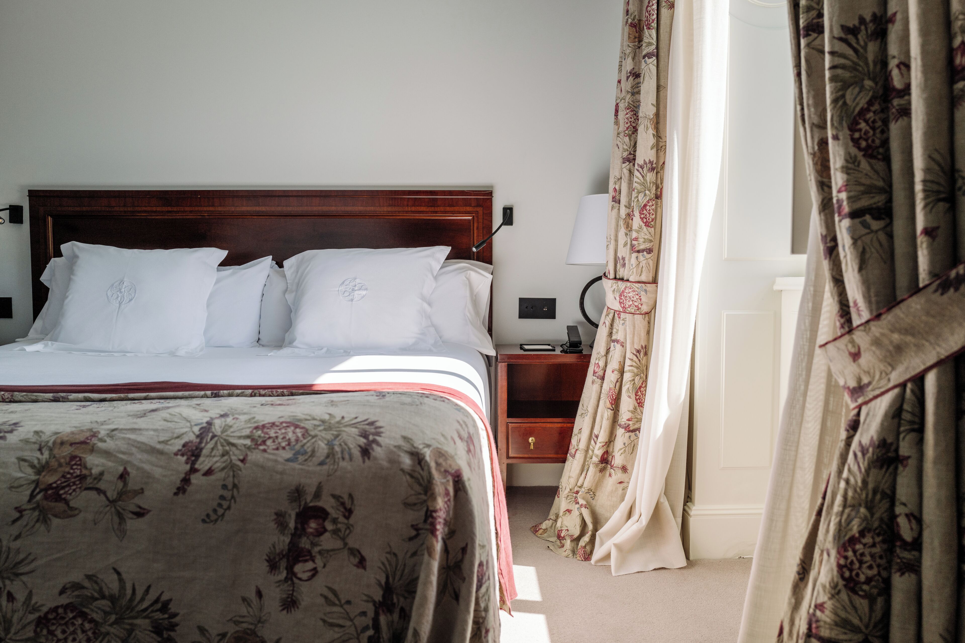 A neatly made bed with a floral bedspread, a wooden headboard, a bedside table with a lamp, and a nightstand with a phone.