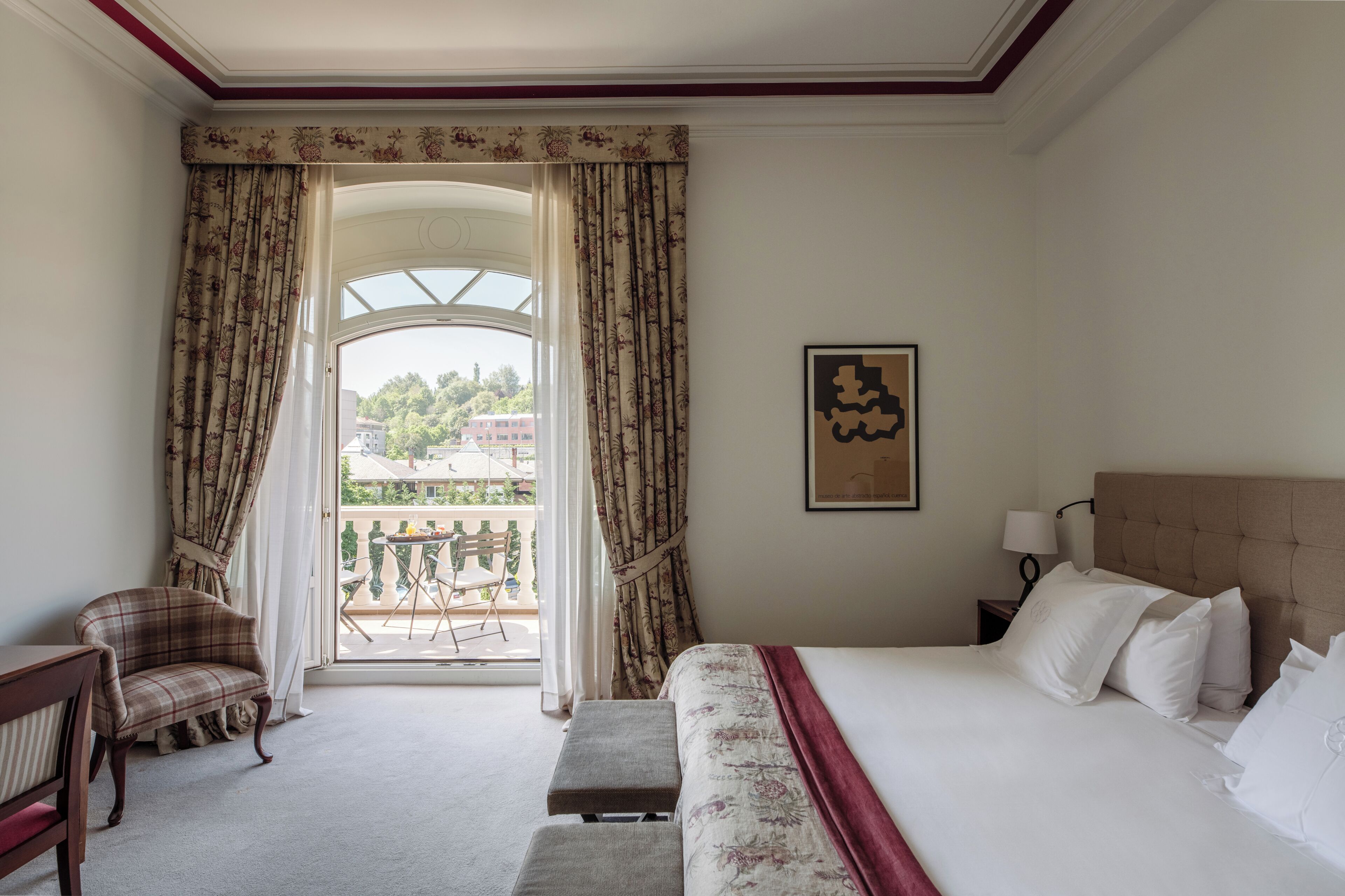 A hotel room with a bed, a chair, a bench, a nightstand, a lamp, a window with curtains, and a balcony with a table and chairs.