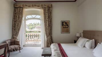 A hotel room with a bed, a chair, a bench, a nightstand, a lamp, a window with curtains, and a balcony with a table and chairs.