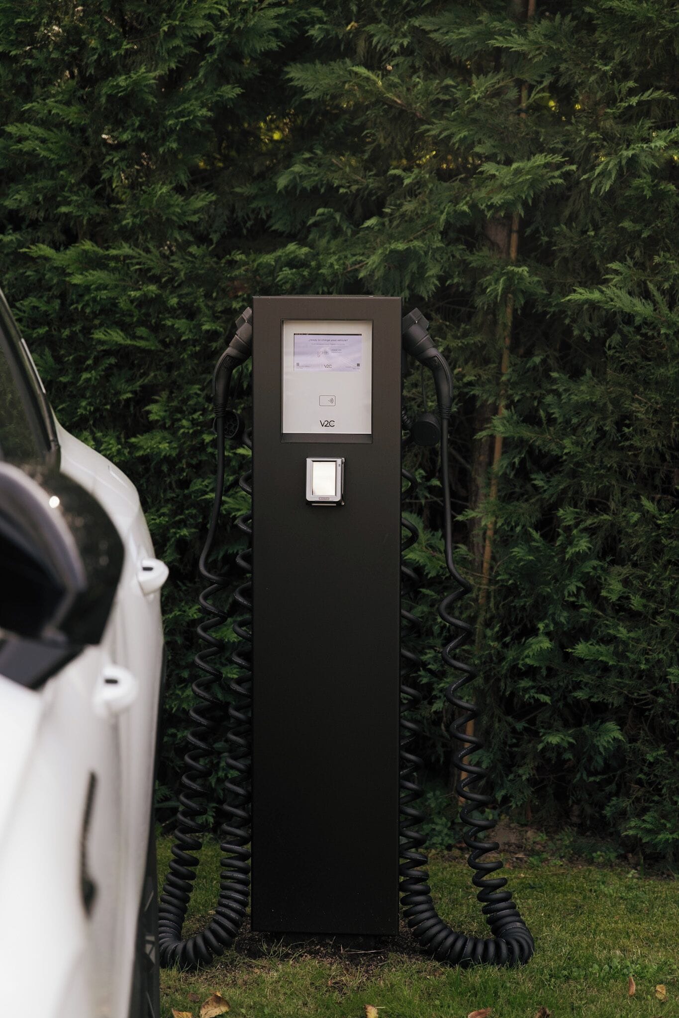 Eine schwarze Ladesäule für Elektrofahrzeuge mit digitalem Display, umgeben von Grünpflanzen.