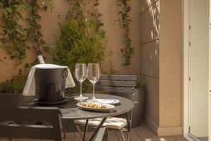 Una mesa preparada con una parrilla cubierta, dos copas de vino y un plato de comida en un patio al aire libre con un banco y follaje de fondo.
