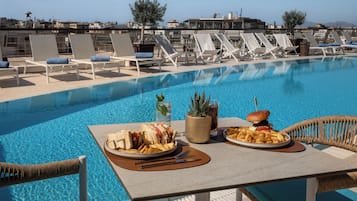Un espace repas aménagé au bord de la piscine, avec une table dressée pour deux personnes, un sandwich, des frites et un hamburger, entouré de chaises longues et offrant une vue dégagée sur le paysage urbain.
