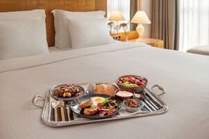 Uma cama bem arrumada com roupa de cama branca, um tabuleiro com itens de café da manhã, incluindo torradas, ovos e frutas, e uma mesa de cabeceira com luminárias.