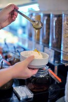 A hand holding a bowl with a spoonful of a white creamy substance, possibly ice cream, in a setting with a jar and a metal container in the background.