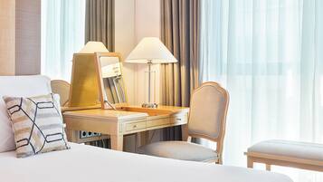 A hotel room with a large bed, a desk with a lamp, a chair, and a view of the outdoors through sheer curtains.