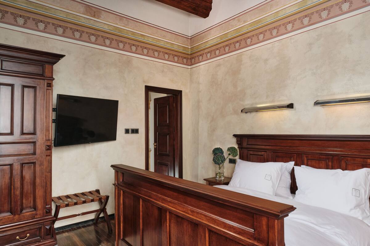 A bedroom with a wooden bed, a television mounted on the wall, a nightstand with a vase of flowers, and a wooden bench.