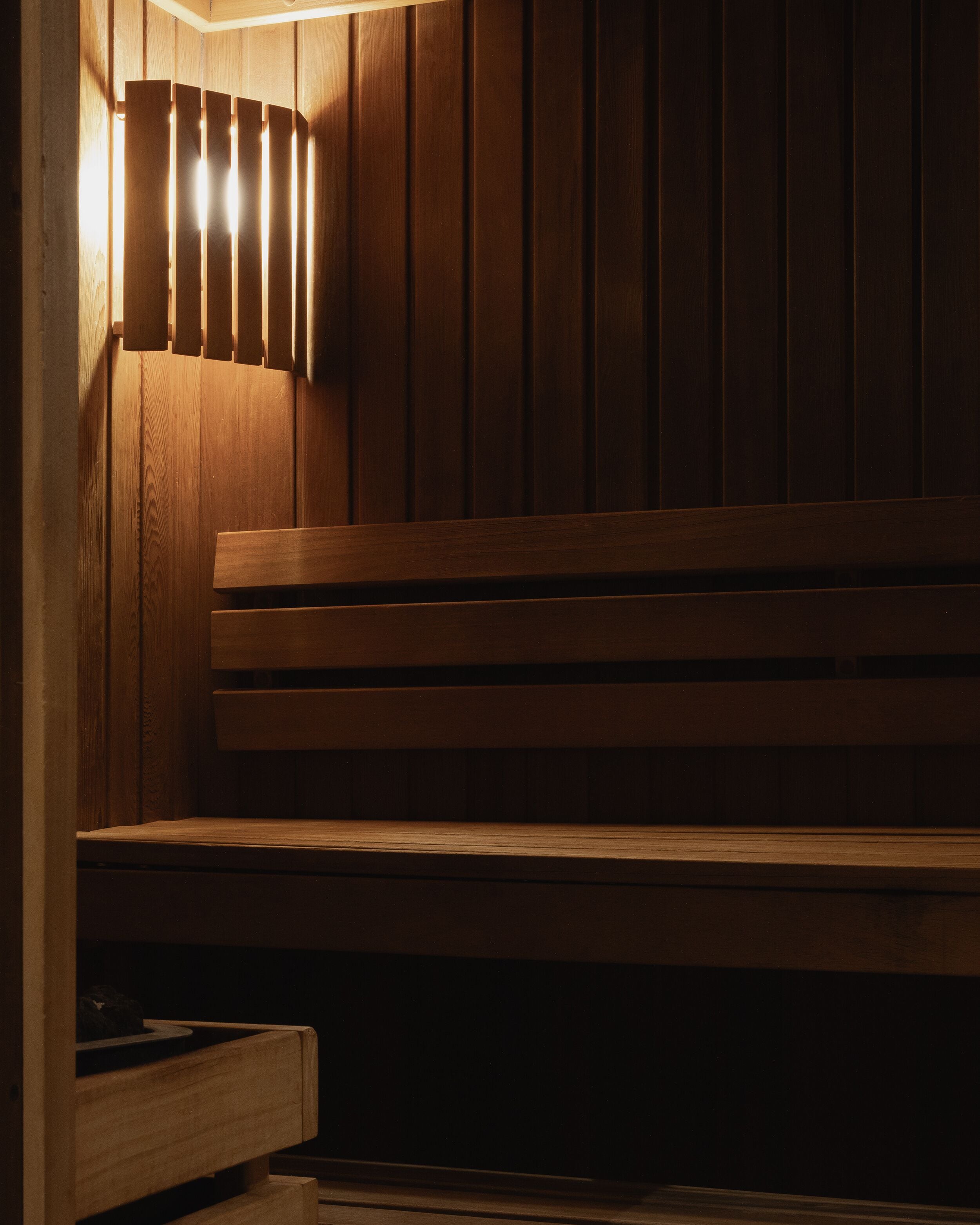 A wooden bench with a slatted backrest and a lower shelf, positioned against a wooden wall with vertical paneling.