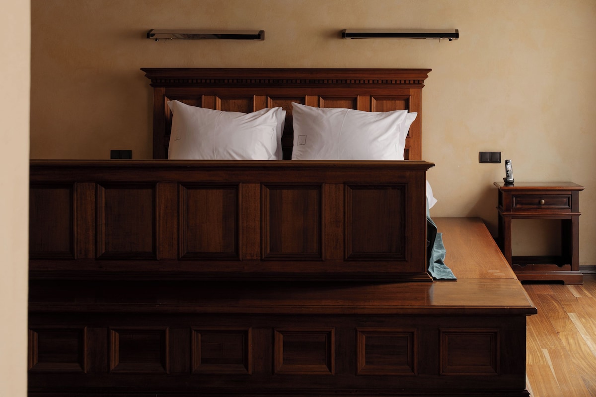 Una cama de madera con cabecera, dos almohadas y una mesita de noche con una lámpara.
