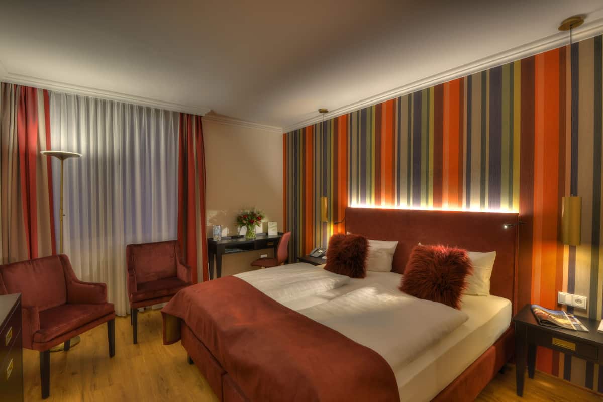 A hotel room with a bed, two chairs, a desk, and a lamp.