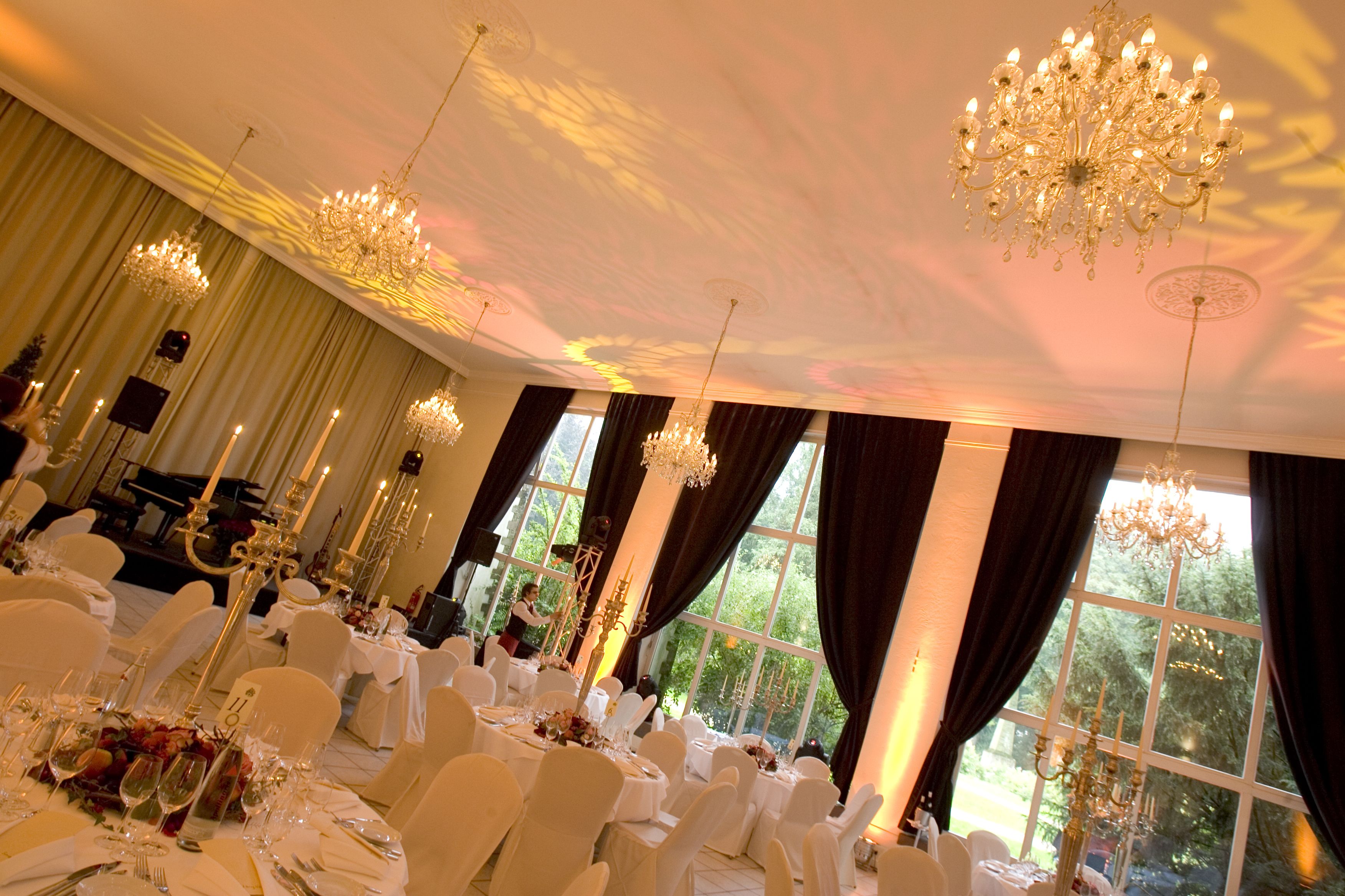 Un salón de banquetes con mesas dispuestas para un evento formal, candelabros y grandes ventanales con cortinas.