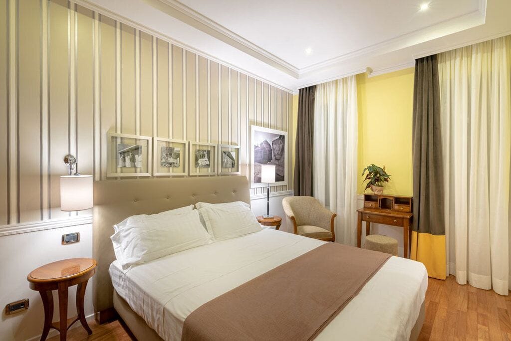 Quarto de hotel com uma cama grande, uma mesa de cabeceira, uma cadeira, uma pequena mesa com uma planta e uma parede com quadros.