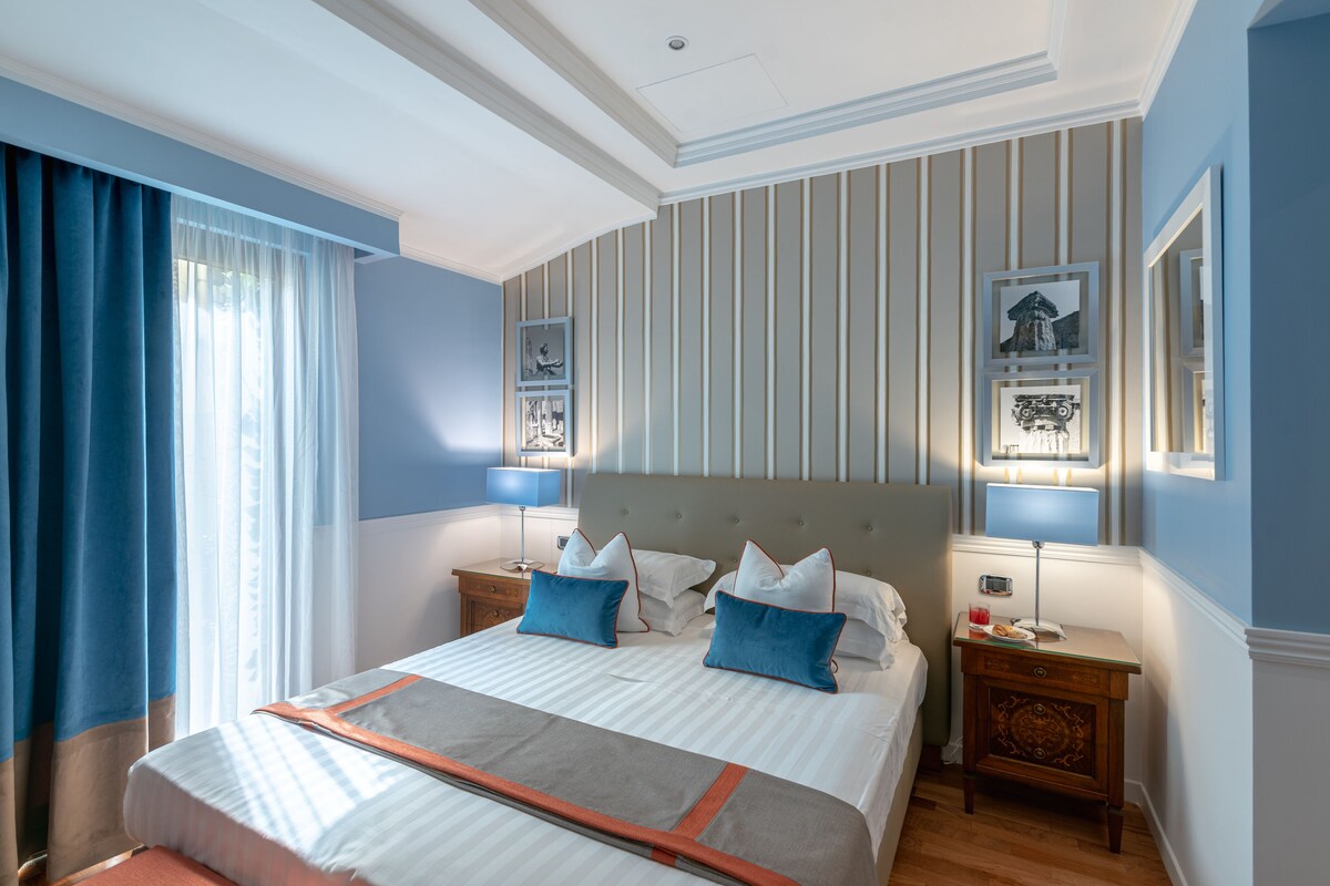 A hotel room with a large bed, two bedside tables, a nightstand, a lamp, and a window with curtains.