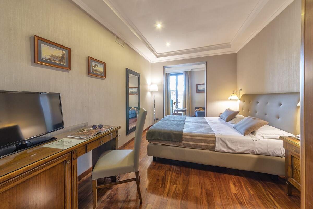 A hotel room with a bed, a desk with a TV, a chair, a bedside table, and a lamp.