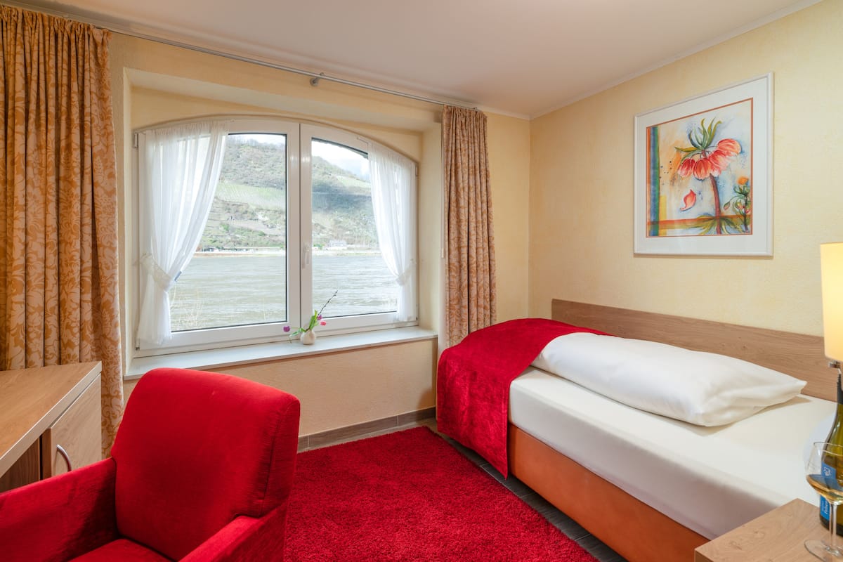 A hotel room with a bed, a desk, a chair, a window with curtains, and a painting on the wall.