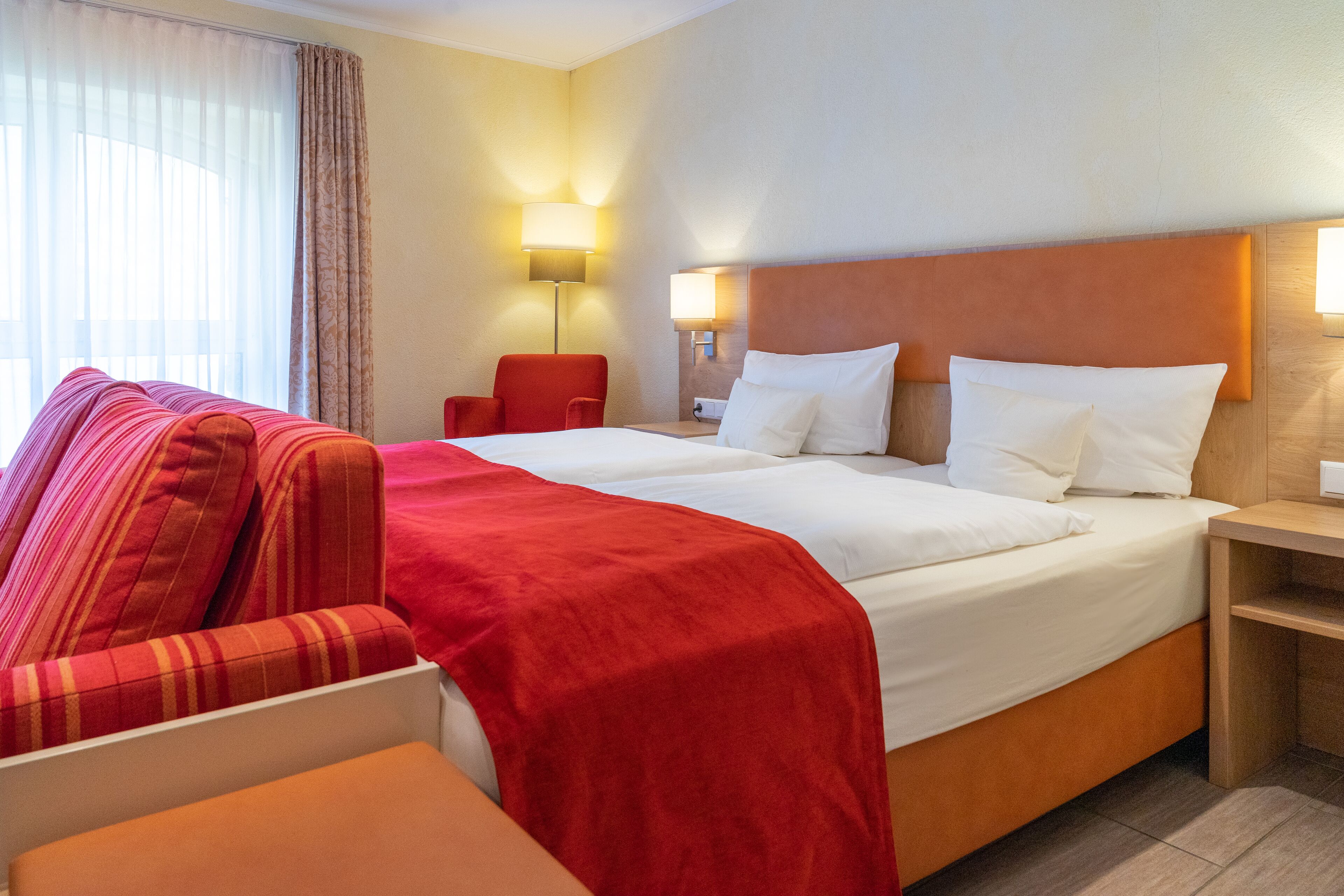 A hotel room with a large bed, a red armchair, a wooden nightstand, and two bedside lamps.
