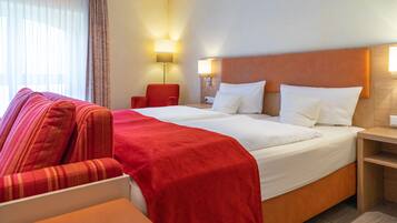 A hotel room with a large bed, a red armchair, a wooden nightstand, and two bedside lamps.