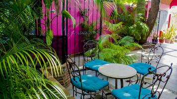 Un patio con una mesa redonda y cuatro sillas azules con cojines, rodeado de exuberante follaje y un muro rosa.