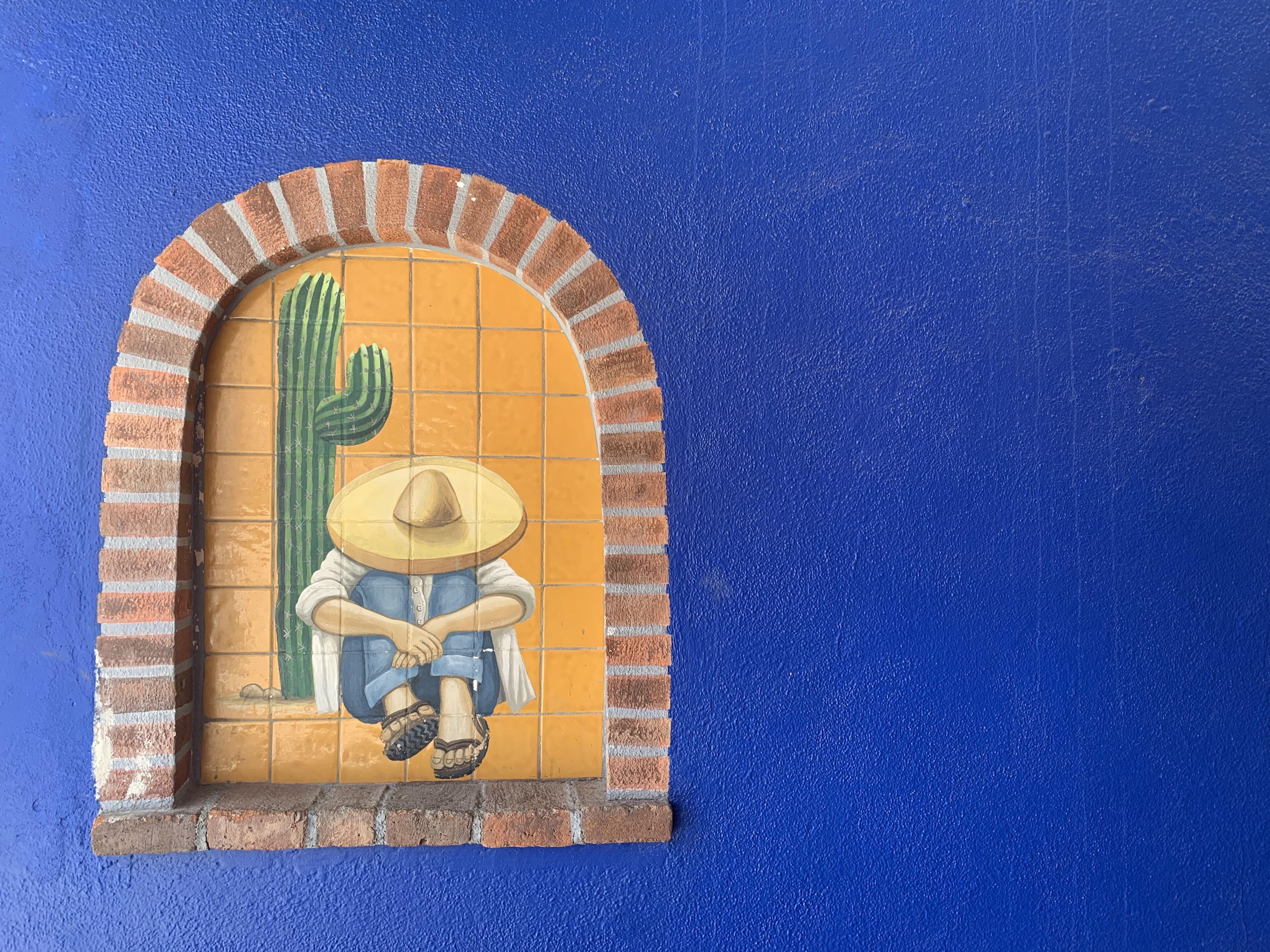 Een muurschildering van een persoon in traditionele kleding, zittend op een bankje, omlijst door een bakstenen boog met een cactus.
