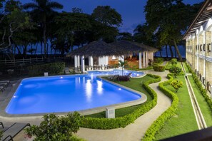 A swimming pool with a thatched-roof structure, surrounded by well-maintained greenery and a clear view of the ocean. - Fuego del Sol Beachfront Hotel (Jacó)