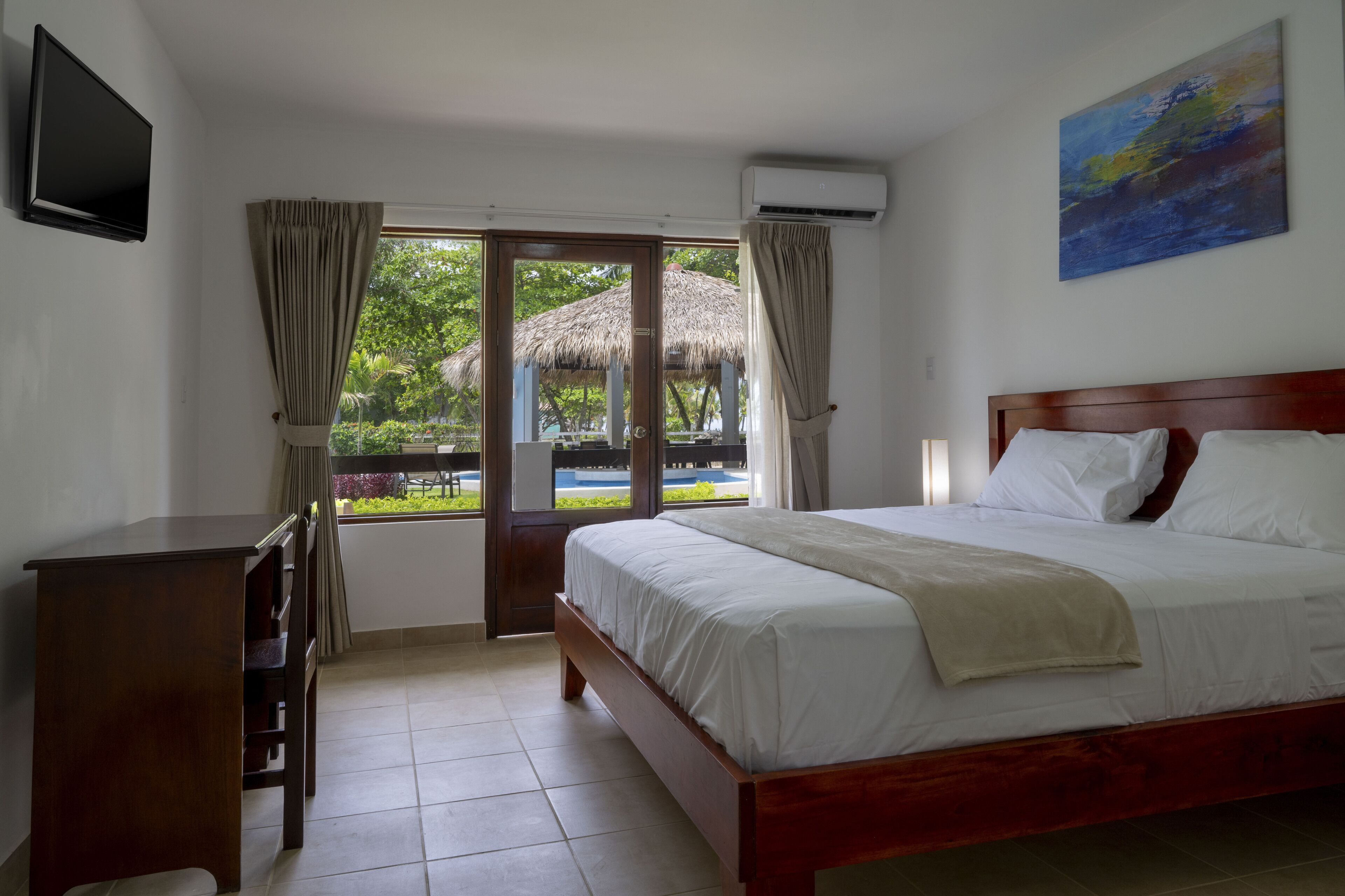 A bedroom with a bed, a desk, a TV, and a view of a thatched structure and greenery.