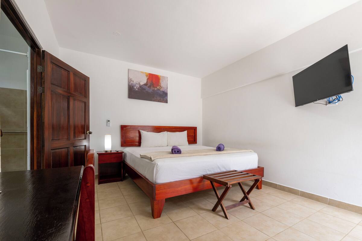 A bedroom with a wooden bed, a bedside table, a TV mounted on the wall, and a painting above the bed.